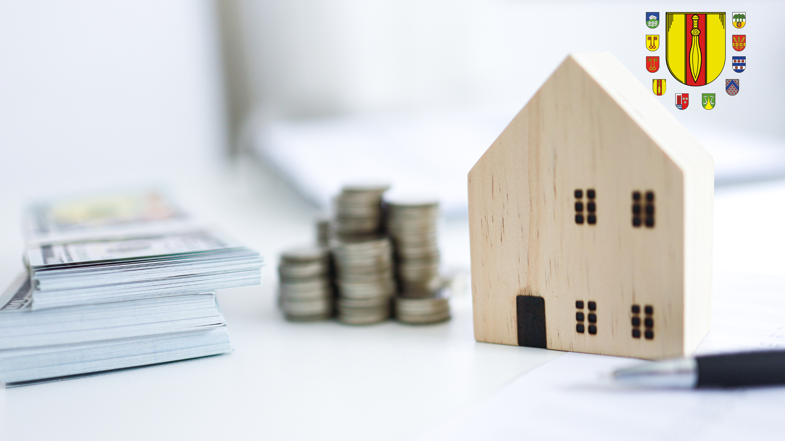 Symbolbild mit Münzen, Geldscheinen, Holzhaus und einem Kugelschreiber.